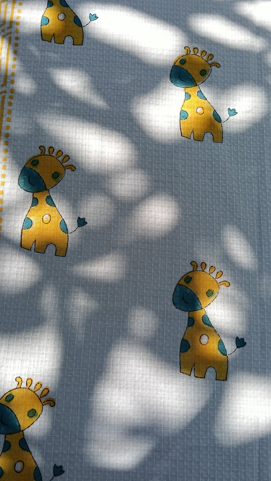 A white cotton bath towel with a hand block print design featuring giraffes in yellow and blue colors.